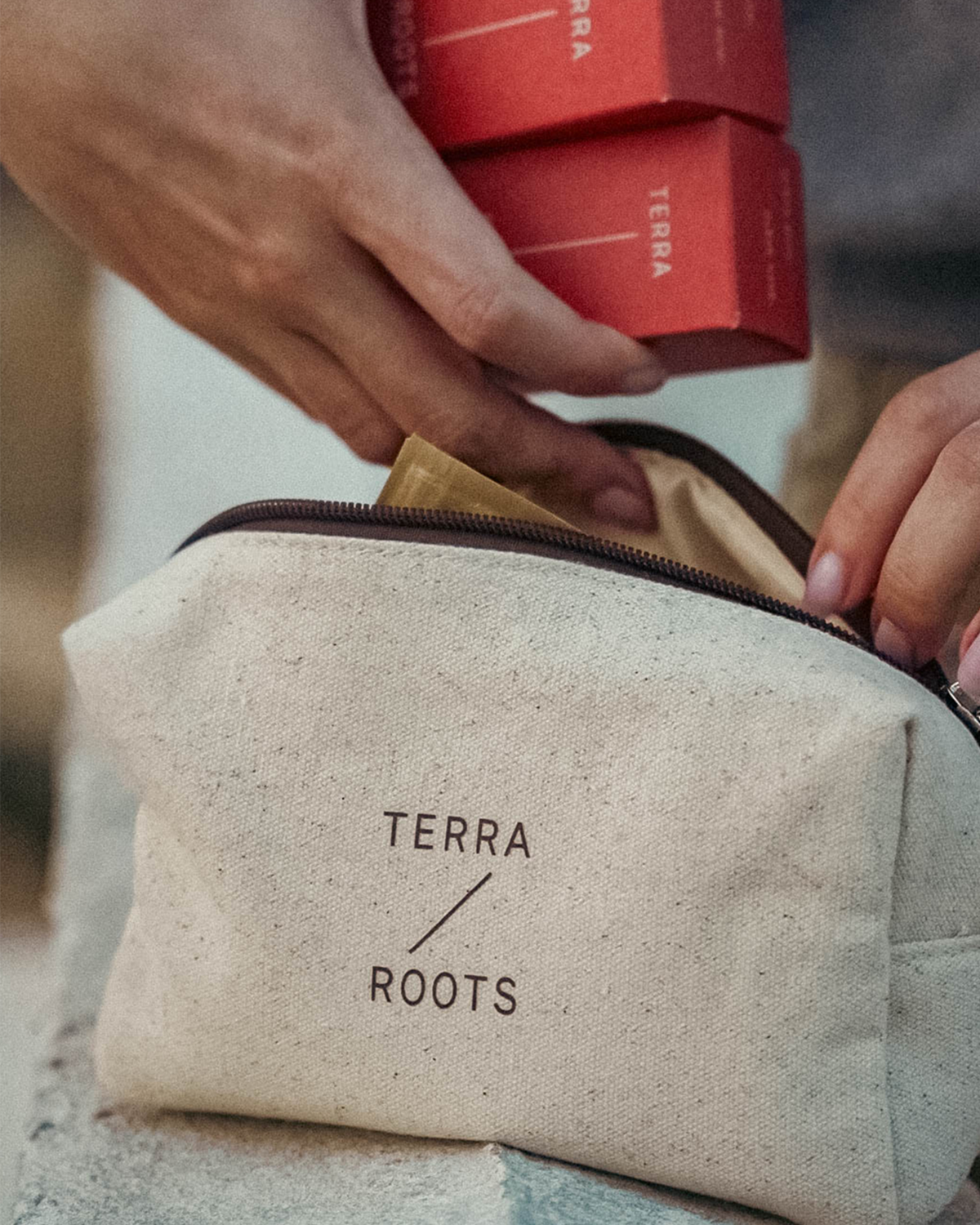 A close-up lifestyle shot of a person's hands placing a gold packet into a beige canvas zippered pouch with black 'TERRA ROOTS' branding. Two red Terra Roots boxes are held above the pouch.